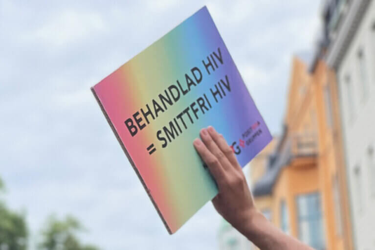 En hand håller upp en regnbårgsfärgad skylt mot himlen. På skylten står det "behandlad hiv = smittfri hiv".
