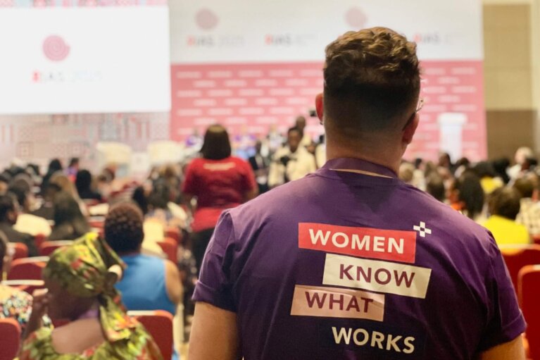 Bild på ryggtavlan på person med en Women Know What Works-tröja som tittar mot en scen på IAS2025.